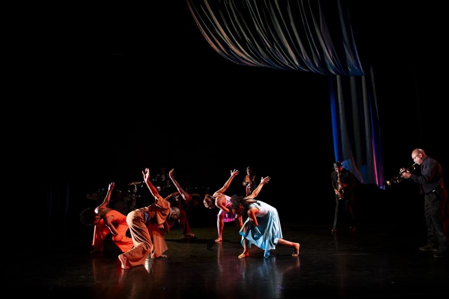 On a stage, with a curtain draped on the right, four Black dancers in bright dresses and big pants lean towards each other and throw their arms back.