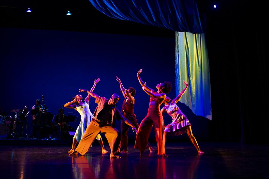On a stage, five Black dancers pose with their arms up, in front of a live band.