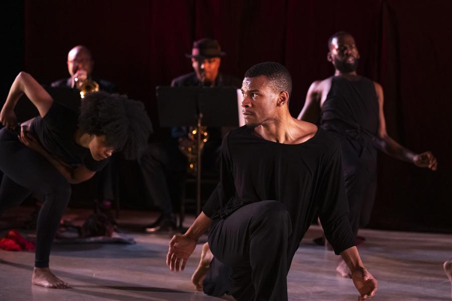 A Black person looks to their left, on one knee. Behind them, three dancers, also in black, dance.