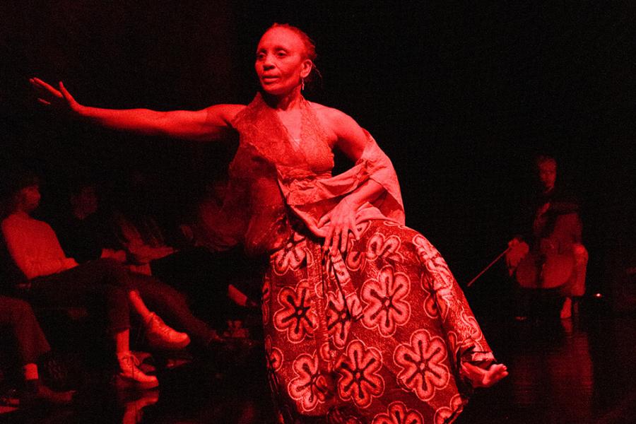 On stage and in red light, a woman holds her dress up, while others dance around her.