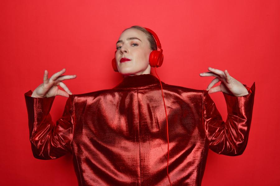 A blonde, white lady poses in all red in front of a red backdrop, including red headphones.