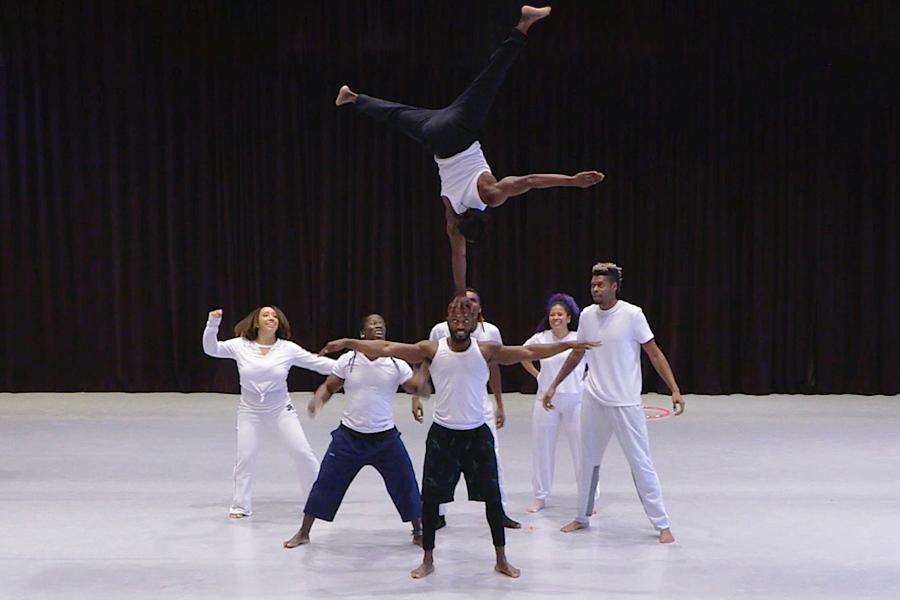 Six Black performers on a white stage. A man, in a white beater, does a one arm stand on the shoulder of one of his peers who has his arms out to balance himself.