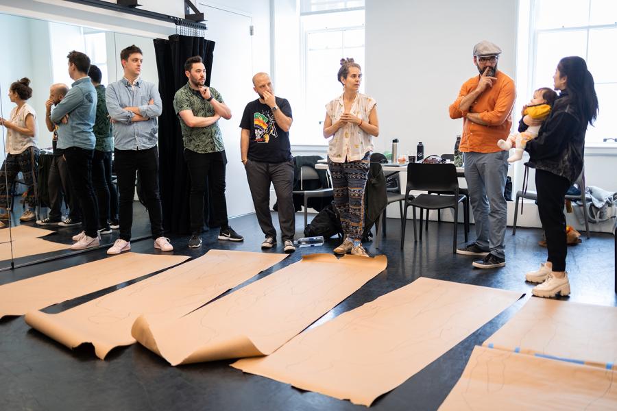Six ensemble members of The Magic Bullet stand in a line in a light-filled rehearsal room with large brown rolls of paper unfurled on the floor in front of them. The ensemble members stand in various attitudes of listening and contemplation as they direct their attention to Caitlin at the end, who is holding an infant wearing a bright yellow shirt. 