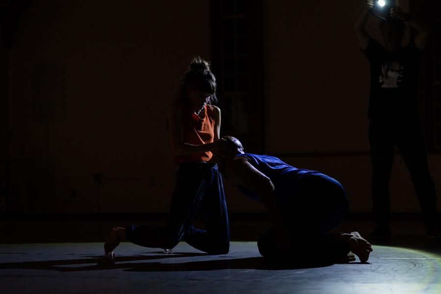 In low light, a white woman touches the bald head of a man who bows before her.