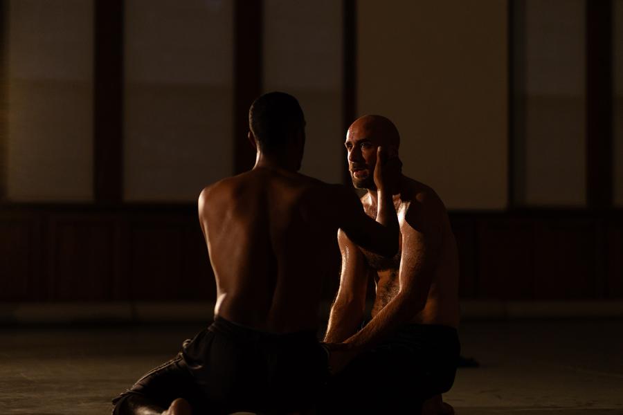 In low light, a shirtless Black man caresses the face of a bald and bearded white man.