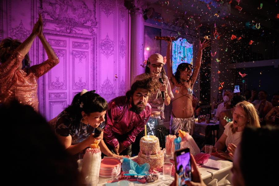 A lively birthday celebration with a group of people gathered around a table at Bowery Poetry Club. In the center, a man and a woman are blowing out candles on a colorful cake while others cheer and celebrate. The scene is vibrant, with confetti flying in the air and party decorations visible, including pink and blue napkins, plates, and utensils. The background features an ornate, pink-lit wall with detailed patterns, adding to the festive atmosphere. Liz and Bow are holding microphones.