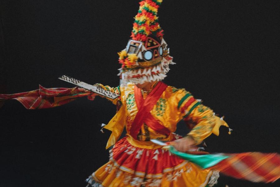 A dancer twirls in an ornate orange, red, and green stripped costume with tassels over their face.