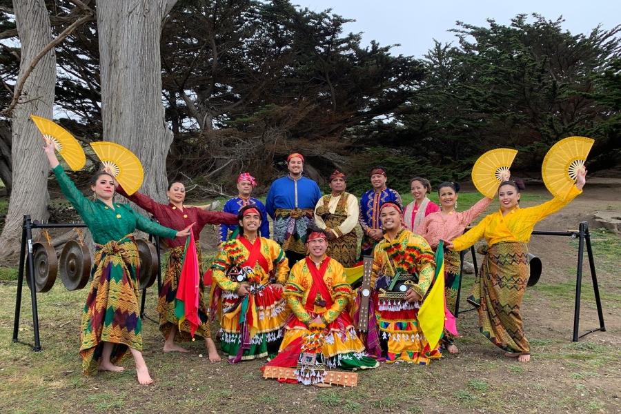 A group poses in a field, while wearing ornate costumes and four of them hold golden yellow fans.