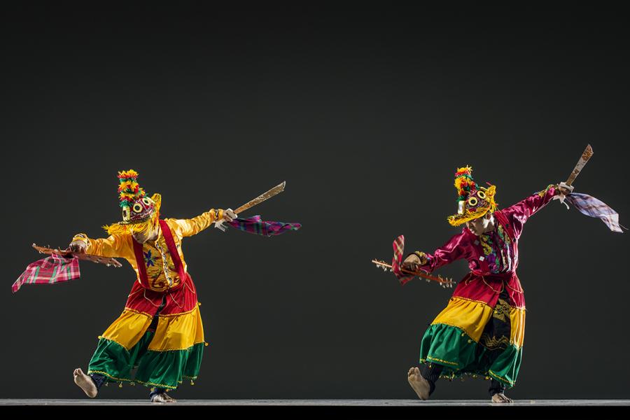 Two dancers in orange, red, and green striped costumes hold their arms out and lean forward.