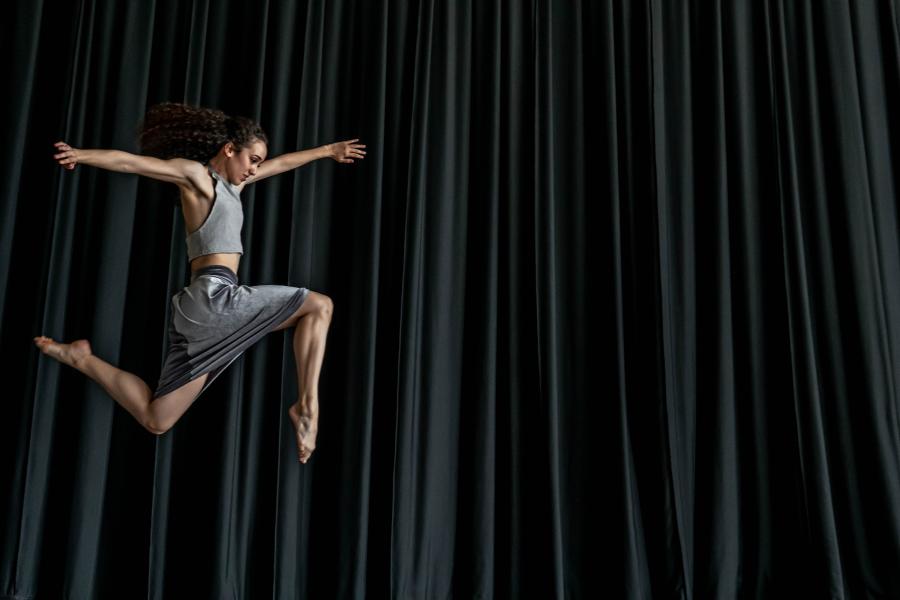 In front of a teal curtain, a dancer, in a grey dress, leaps.