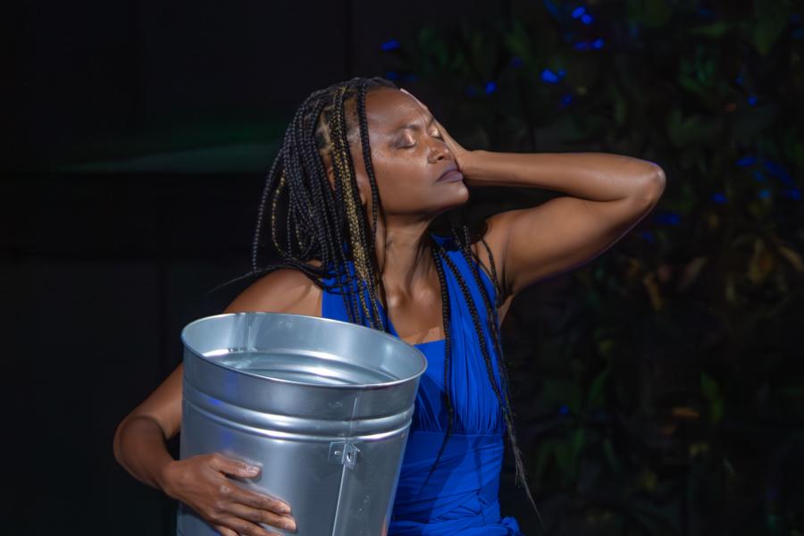 A Black woman, in stage lighting, holds a tin bucket and her forehead. 
