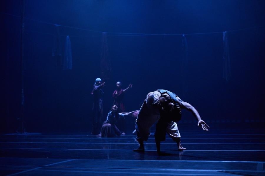 On a dark stage, with moody blue lights, a man leans over another dancer backwards.