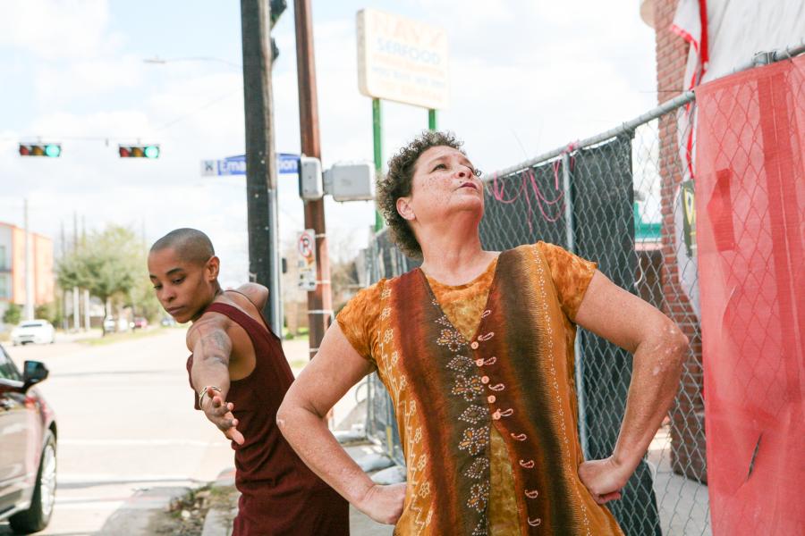 On a city street, two women of color, in green and brown dresses, pose with their hands on their hips and with their arms stretched wide.