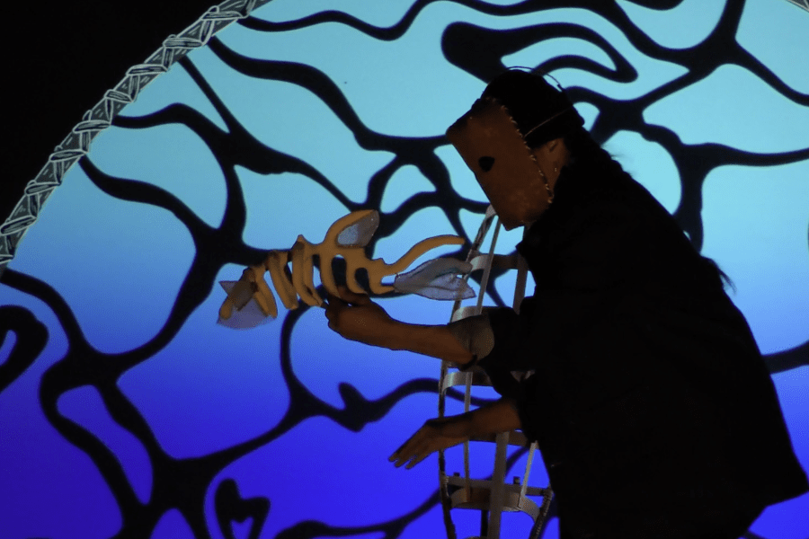 Silhouette of a figure in motion wearing a mask, holding a puppet shaped as a fish, and standing in front of the propped-up skeleton of a birchbark canoe. The illustration behind the figure glows in shades of blue, with waves sketched in black.