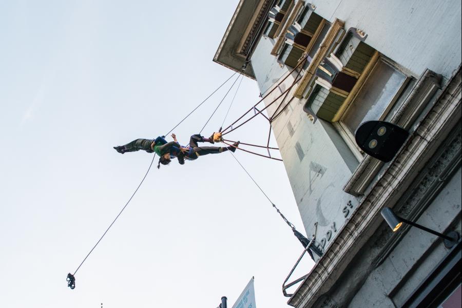 In daylight, two performers dangle from a web of cords off the side of a building.