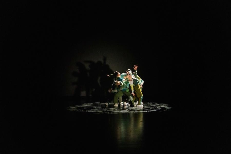 In a dark studio space with a spotlight, dancers lean on each other with their hands out in different directions