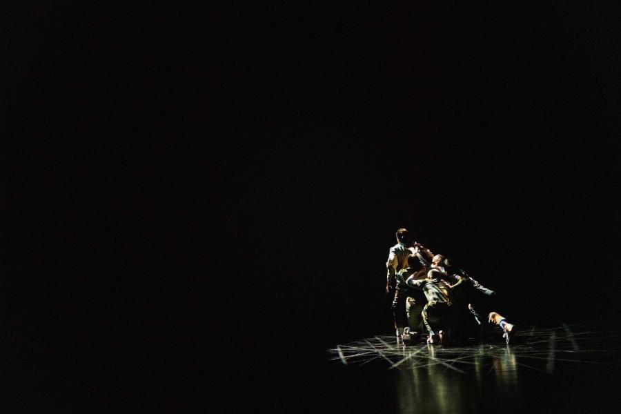 From behind, in a dark studio space, two dancers grab at another who walks away. 