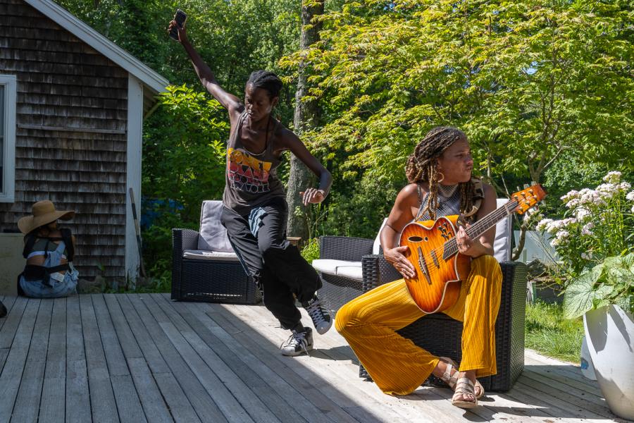 On a porch, a Black man dances while a Black woman, with long braids, plays the guitar.
