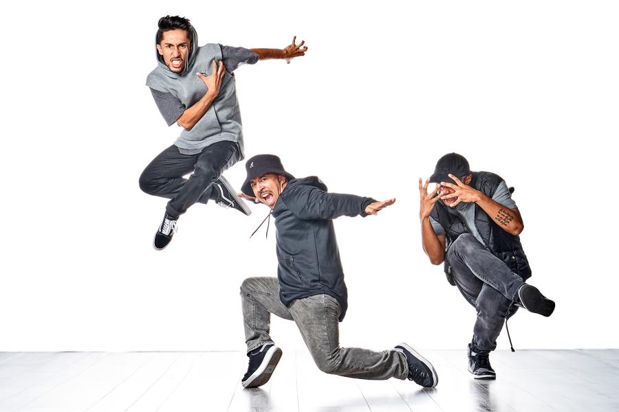 Three dancers wear gray in front of a white backdrop. One leaps with his arms thrown back. Another slides and smirks. Another crosses his legs and covers his face with his fingers spread.