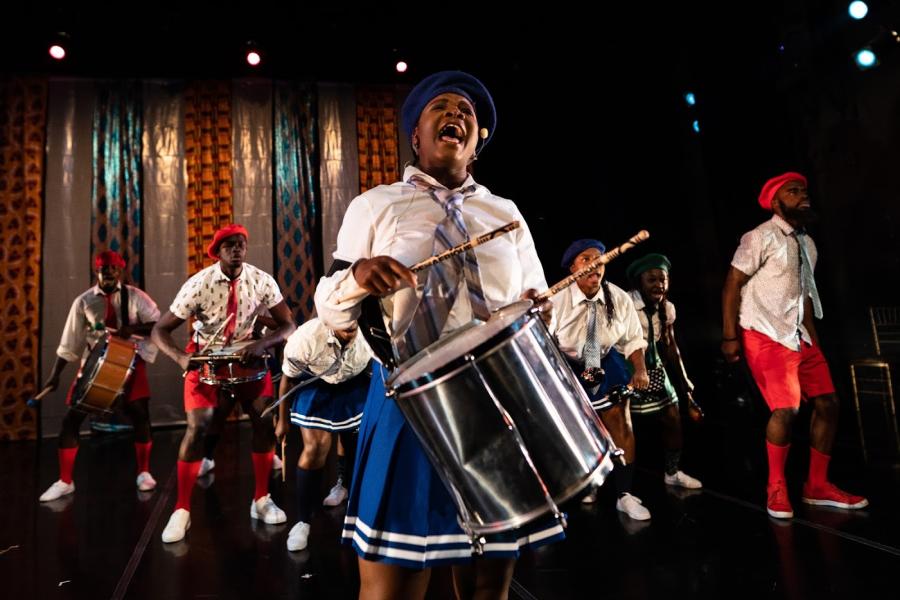 A Black girl plays the drums and sings. Behind her six more folks sing, dance, and play music in red and blue uniforms.
