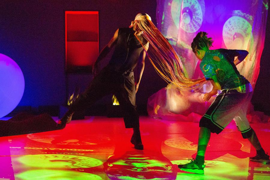 In a gallery, with projections of different colors, two dancers swap. One has a long mask on that looks like fringe.