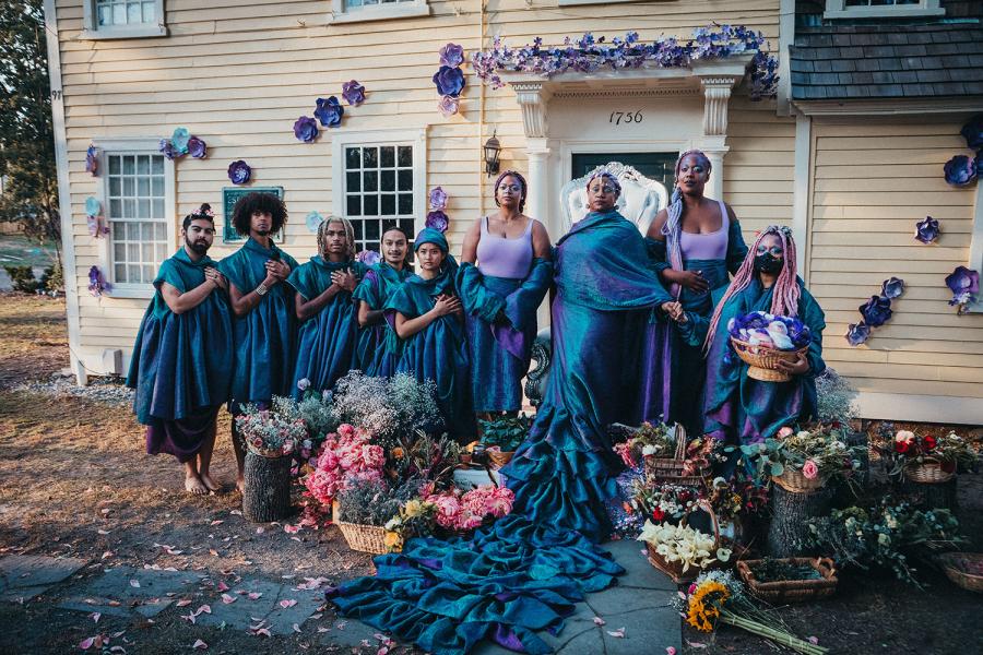Nine folks, in blue gowns and purple tank tops, pose in front of a yellow house that has been decorated with flowers. In the center, one of the people has a veil and a long trail on their blueish gown.