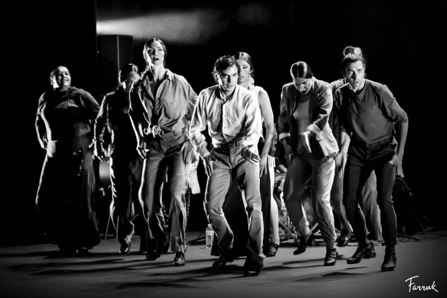 In black and white, a group of folks, in button-downs and casual dress, are lit dramatically and crouch forward.