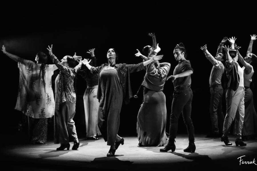 In black and white, a group of dancers throw their arms up in the air and hop on one foot.