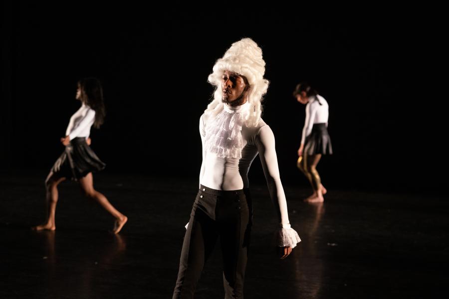 A black man in a tall white colonial style wig performs in front of two female dancers.