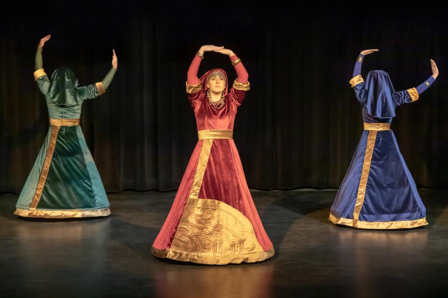 On a black stage with black curtains, three dancers wear dresses with gold accents. One faces the audience in red, two others in green and blue have their backs to us.s 
