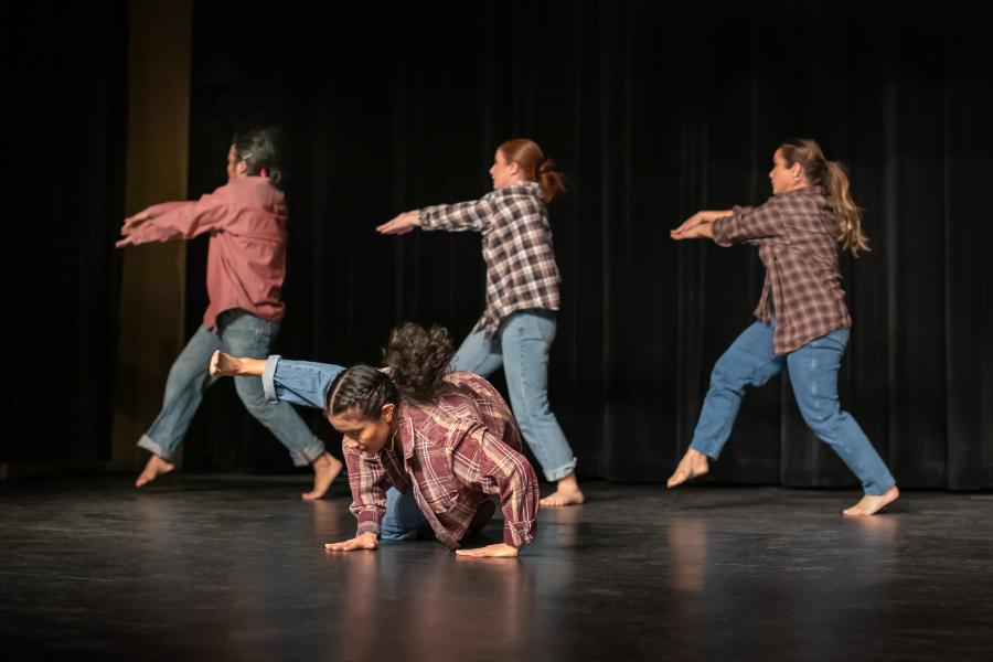 Four dancers, on a black stage with black curtains, wear denim and flannel. Three face left and throw their arms that way; another is on the ground kicking in the same direction.