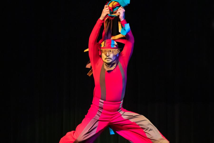 On a black stage with black curtains, a dancer leaps in a jester costume.
