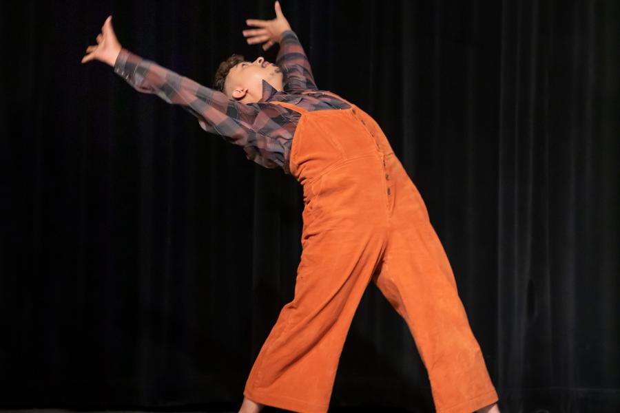 On a black stage with black curtains, a dancer, in orange overalls, leans backwards.