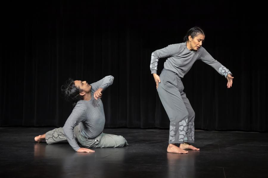 On a black stage with black curtains, two dancers, with jet black hair, wear gray sweatsuits.