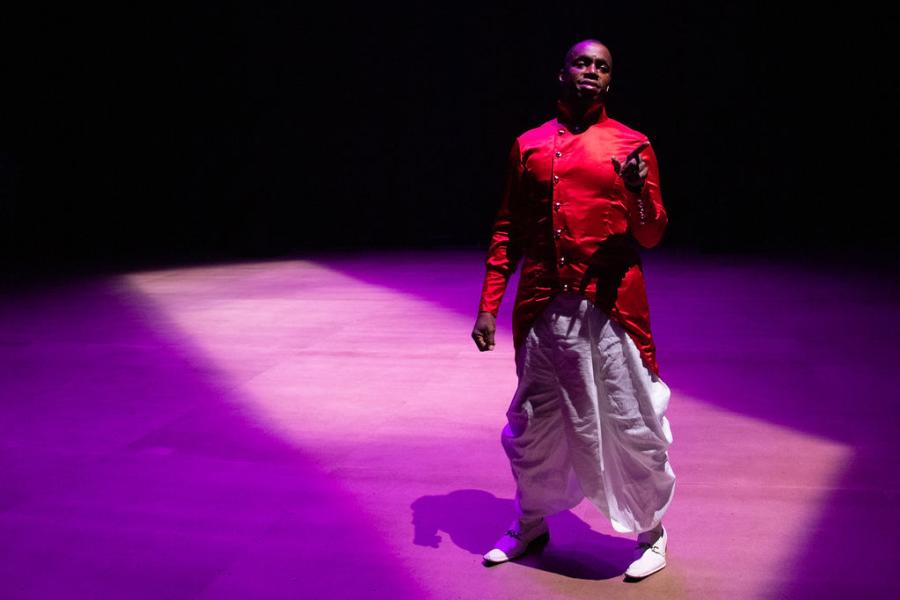 A Black man wears a red trench with coat tails and baggy white pants. He's in a spotlight on a stage that is lit in purple.