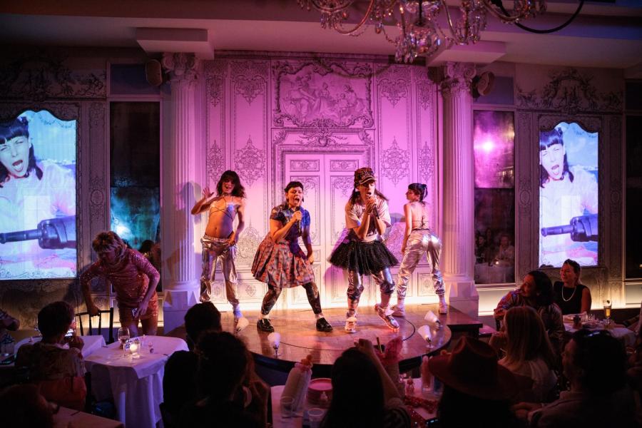 A performance on a small stage with five performers in colorful and eclectic costumes. The two central performers, Liz and Bow, hold a microphone, one wearing a dress and the other wearing a tutu over pants, while others around them are dressed in shiny, silver pants and bikini star shaped tops. One performer off stage is in drag, wearing a sequin pink dress. The background is an ornate, pink-lit wall with detailed patterns, enhancing the dramatic and artistic atmosphere. 