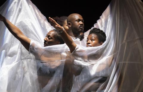In a spotlight, three Black folks hold their arms up. A white sheer fabric is draped over them.