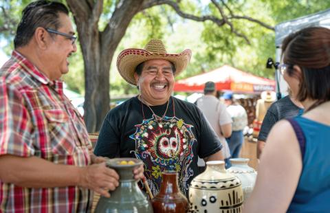 A man in a sombrero holds vases with his friend. A white lady stands on the other side of their table.