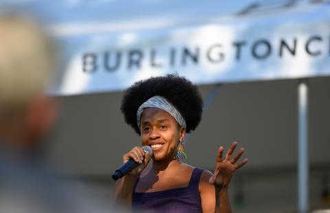 A Black woman, with a short afro, speaks into a microphone. Behind her a sign reads "Burlington."