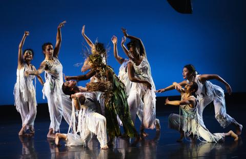 On a stage with a blue background, dancers learn over and toward a dancer who kneels and leans backwards.