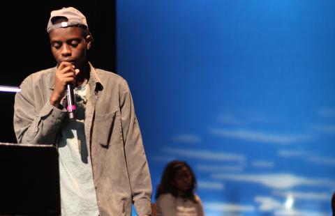 Newcomer Play Project performance photo. Man speaking at a microphone. Photo by Lucy Gellman.