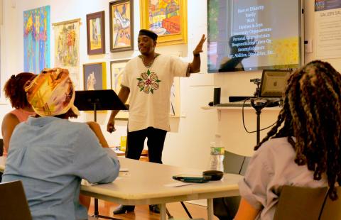 In a classroom, a Black man speaks and points at a screen with text on it.