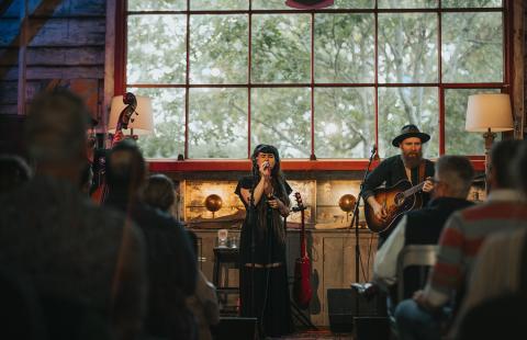 A white lady with dark brown hair sings with her band. The other bandmade we can see has a long beard is also white. The space has a big window with panes. The audience is blurred in the foreground.