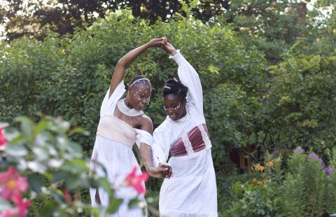 In a garden, two Black women, in white dresses spin with their hands clasped together and their backs to each other.