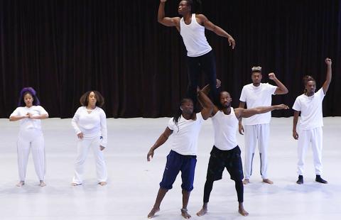Seven Black performers on a white stage. One man, in a wife beater, is on the shoulders of two of his peers and looks out to the left.