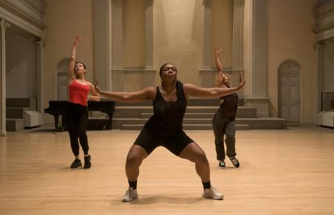 A Black woman, in spanx, holds her arms out in front of two other female dancers.