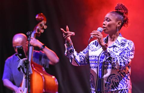 On a stage with red light and fog, a Black woman sings in front of a Black man who plays standing bass.