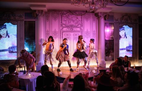 A performance on a small stage with five performers in colorful and eclectic costumes. The two central performers, Liz and Bow, hold a microphone, one wearing a dress and the other wearing a tutu over pants, while others around them are dressed in shiny, silver pants and bikini star shaped tops. One performer off stage is in drag, wearing a sequin pink dress. The background is an ornate, pink-lit wall with detailed patterns, enhancing the dramatic and artistic atmosphere. 