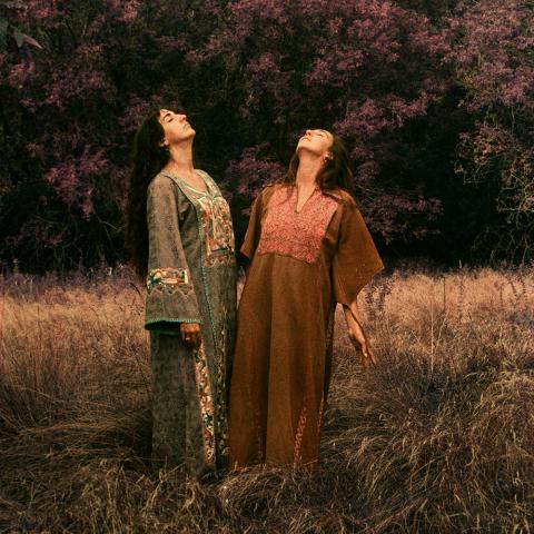 Two women, in long robes, stand and close their eyes. They're in a field.