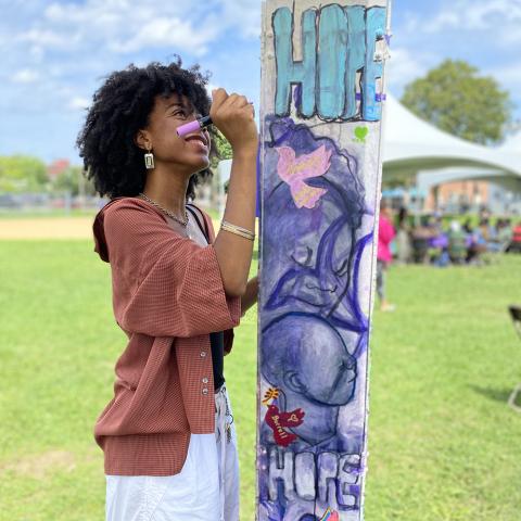 A young black girl, with a afro and a brown, hooded, baseball jacket holds a marker pen to an art installation around a pole.